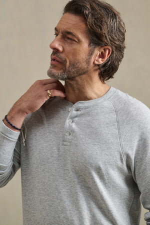 A man in the WESLEY HENLEY by Velvet by Graham & Spencer touches his neck and gazes thoughtfully to the side against a neutral background.