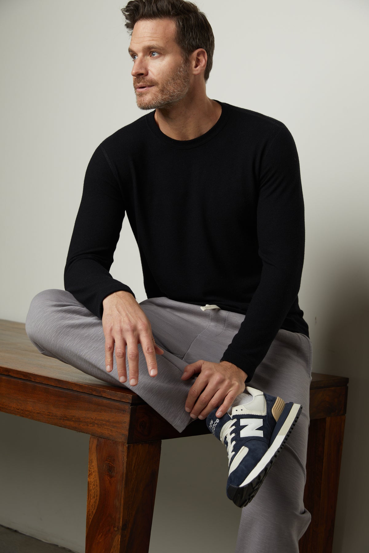   A man sitting on a wooden table wearing a black Velvet by Graham & Spencer sweater and grey Velvet by Graham & Spencer PARKER SWEATPANT pants. 