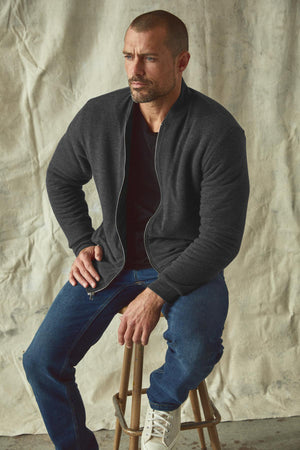 A man sitting on a stool in a cozy Velvet by Graham & Spencer MILES ZIP-UP JACKET and jeans.