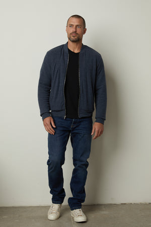 A man is standing in front of a wall wearing MILES ZIP-UP JACKET by Velvet by Graham & Spencer jeans and a casual jacket.