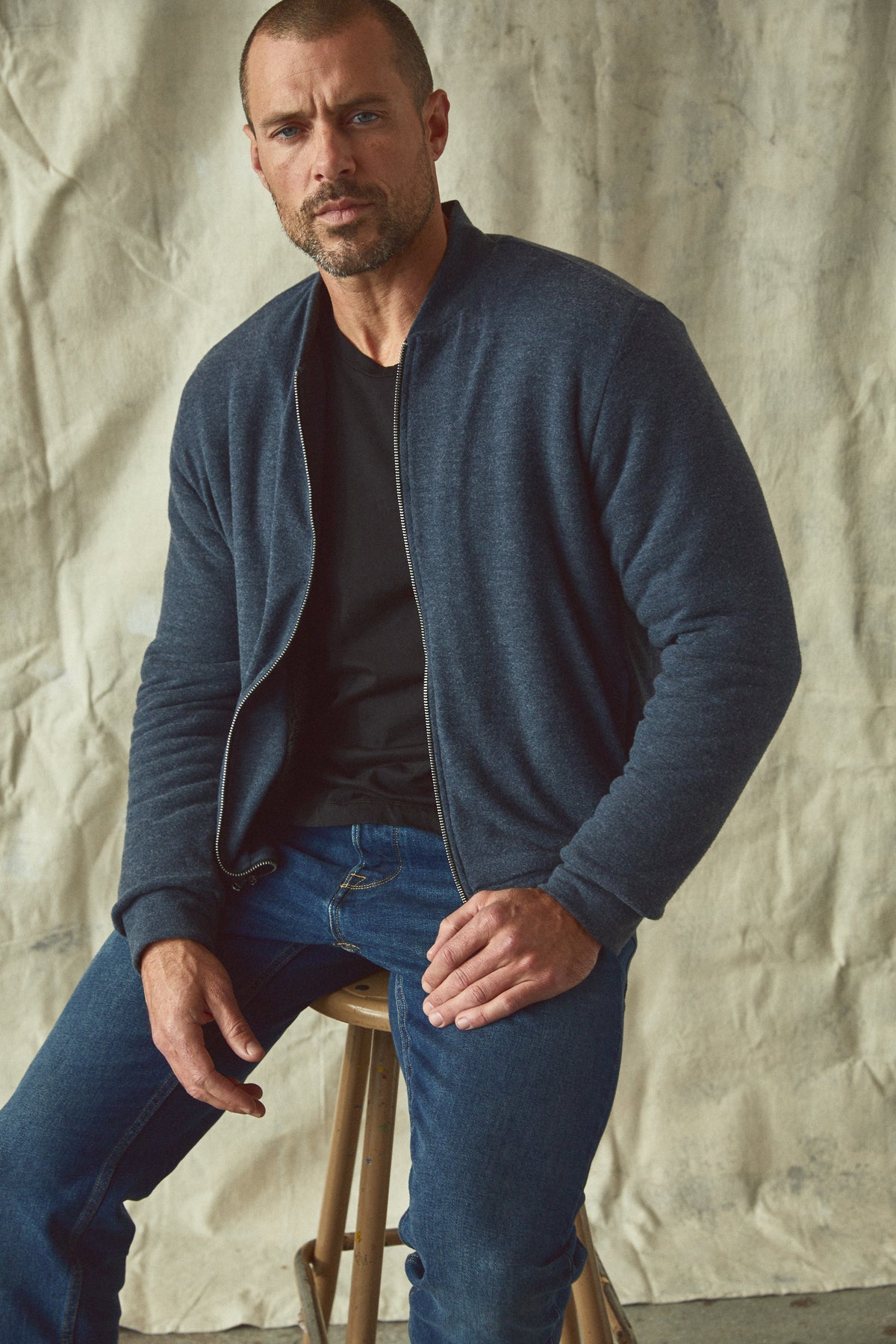 A man sitting on a stool wearing cozy jeans and a Velvet by Graham & Spencer MILES ZIP-UP JACKET.-35662690025665
