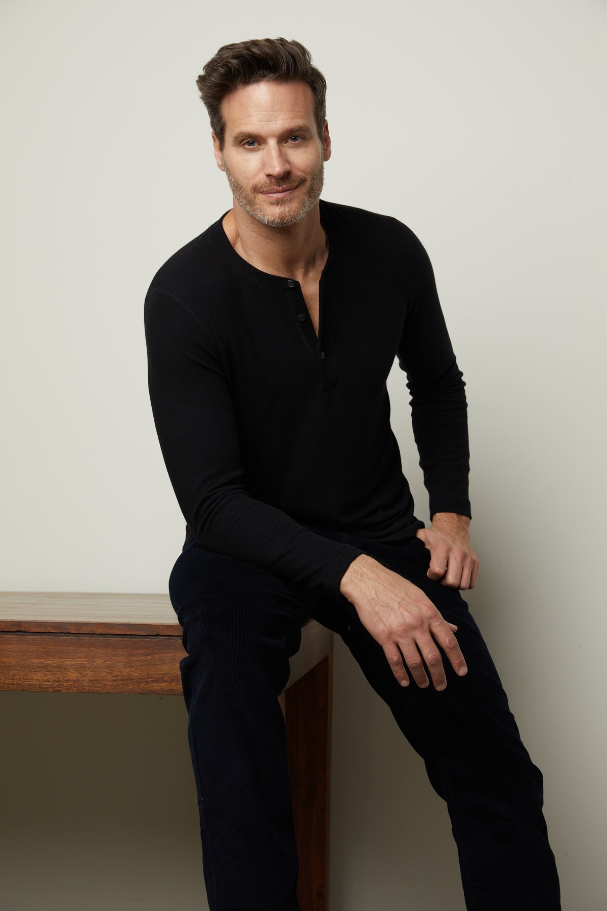   A man sitting on a wooden table wearing a cozy black Velvet by Graham & Spencer Ryland Henley shirt. 