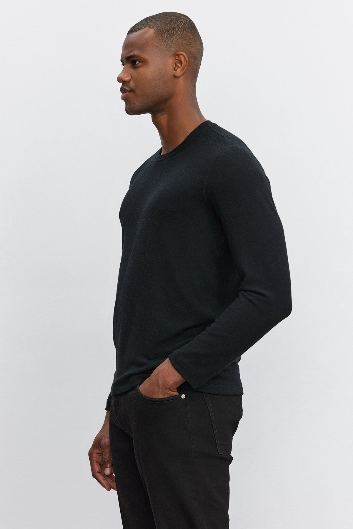   A man with short hair stands in profile against a plain background, wearing the BECKER CREW long-sleeved shirt by Velvet by Graham & Spencer and dark jeans, with one hand in his pocket. 