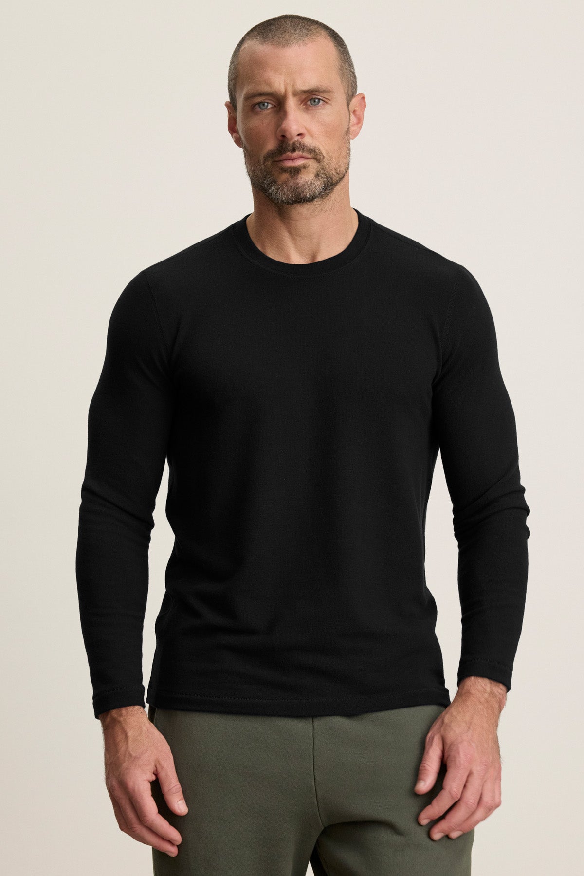 A man with short hair and a beard wears the BECKER CREW by Velvet by Graham & Spencer, a fitted black long-sleeve shirt paired with olive green pants, ideal for daily wear, standing against a plain light background.-40452478861505