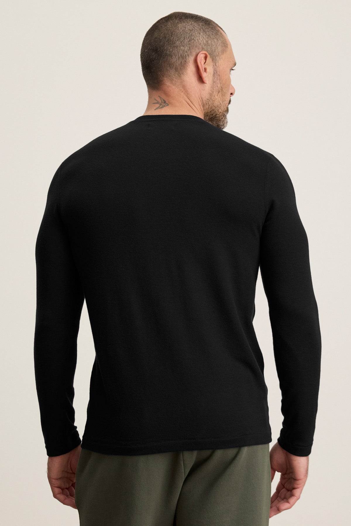 A man with short hair and a neck tattoo is seen from the back wearing the BECKER CREW by Velvet by Graham & Spencer, a cozy black jersey long-sleeve shirt, paired with olive green pants against a plain background.-40452478894273