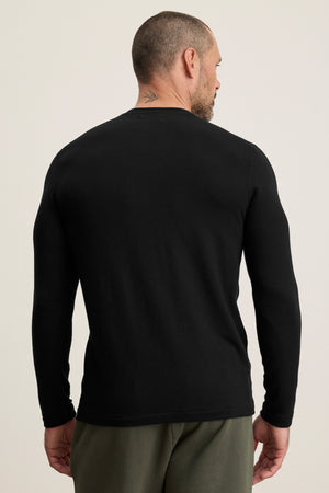 A man with short hair and a neck tattoo is seen from the back wearing the BECKER CREW by Velvet by Graham & Spencer, a cozy black jersey long-sleeve shirt, paired with olive green pants against a plain background.