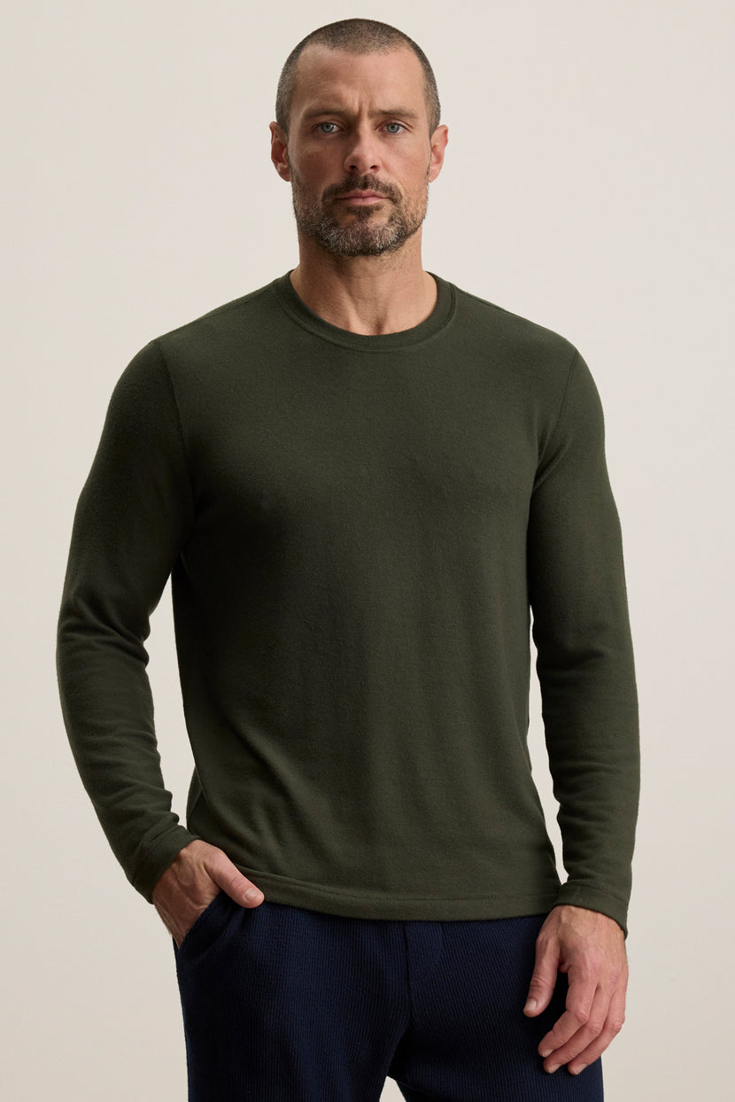 A man with short hair and a beard models the BECKER CREW by Velvet by Graham & Spencer—an olive green fitted long-sleeve crew neck shirt paired with dark pants, standing confidently with his left hand in his pocket against a plain background.