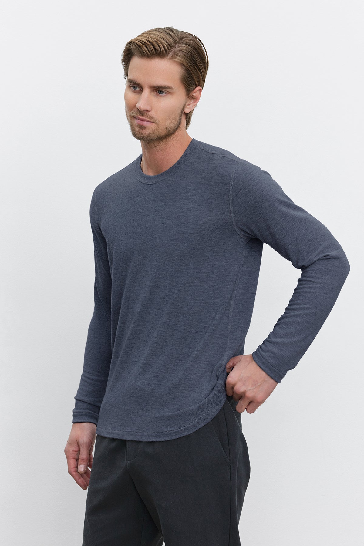   A man with short hair is wearing the BECKER CREW, a fitted long-sleeve shirt in dark gray from Velvet by Graham & Spencer, paired with dark pants. He stands against a plain white background, exemplifying an ideal daily wear style. 