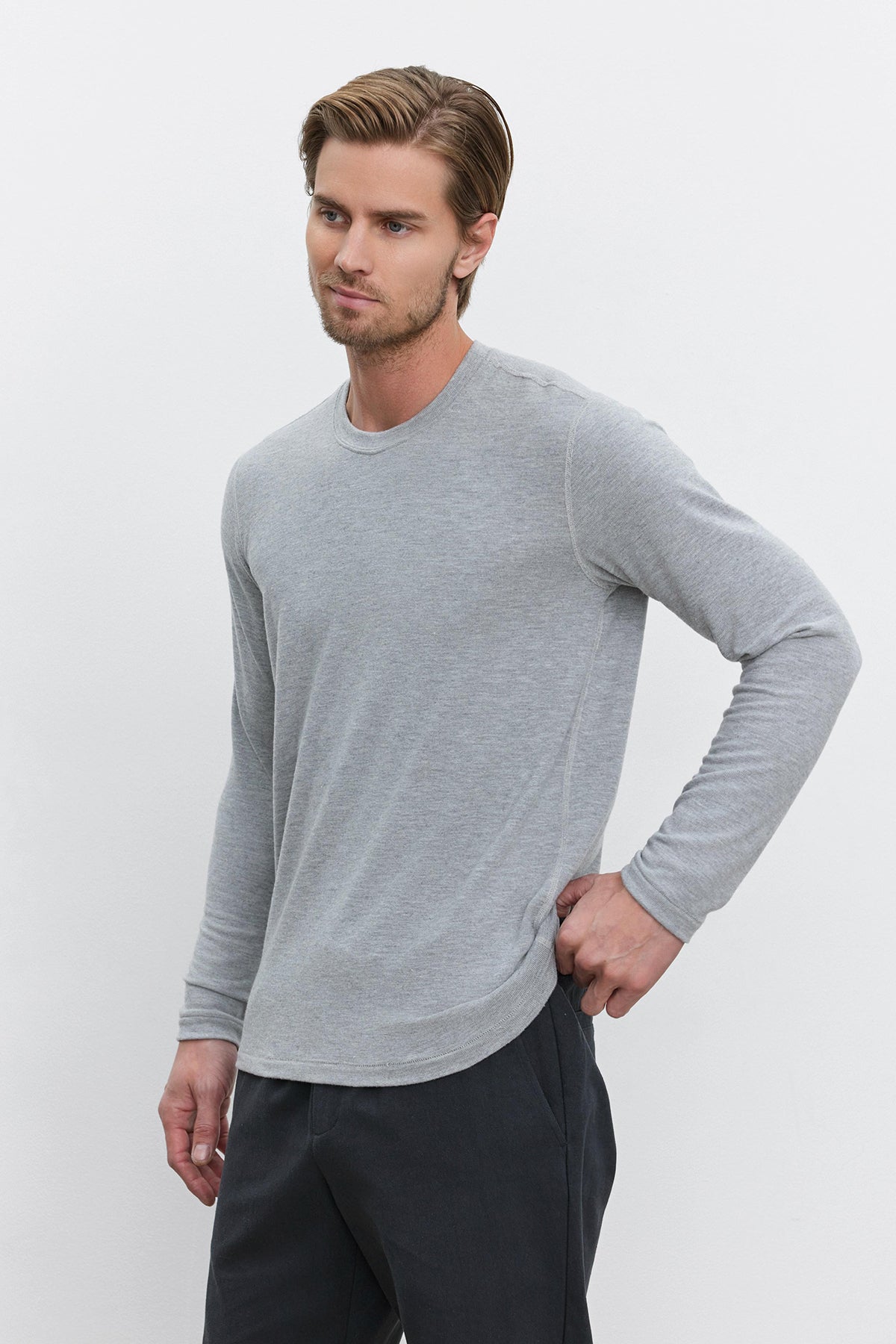   A man in a cozy Velvet by Graham & Spencer BECKER CREW jersey gray long-sleeve shirt and dark pants stands against a plain white background with one hand on his hip. 