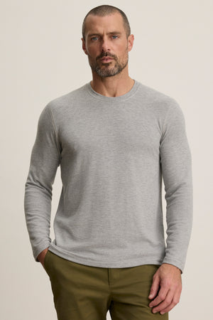 A man with short hair and a beard wears the Velvet by Graham & Spencer BECKER CREW light gray long-sleeve shirt and olive green pants, standing against a plain light background.