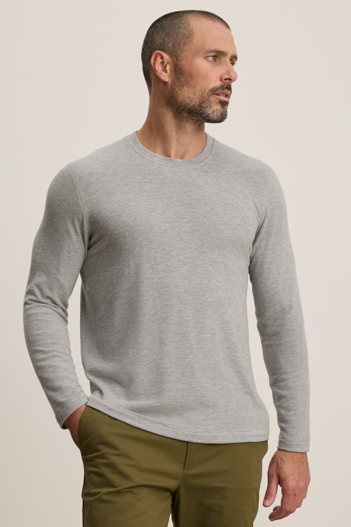 A man with short hair and a beard wears the Velvet by Graham & Spencer BECKER CREW, a cozy light gray fitted long-sleeve crew neck shirt, paired with olive green pants, standing against a plain background and looking left.-39680468844737