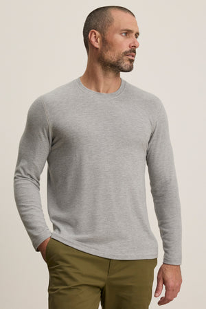A man with short hair and a beard wears the Velvet by Graham & Spencer BECKER CREW, a cozy light gray fitted long-sleeve crew neck shirt, paired with olive green pants, standing against a plain background and looking left.