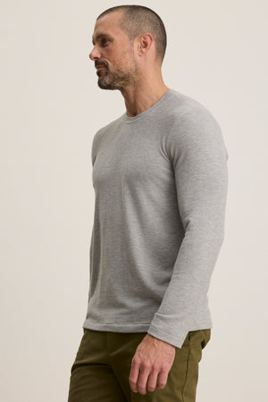 A man with short hair and a beard models the BECKER CREW light grey long-sleeve shirt by Velvet by Graham & Spencer, paired with olive green pants, against a plain background.