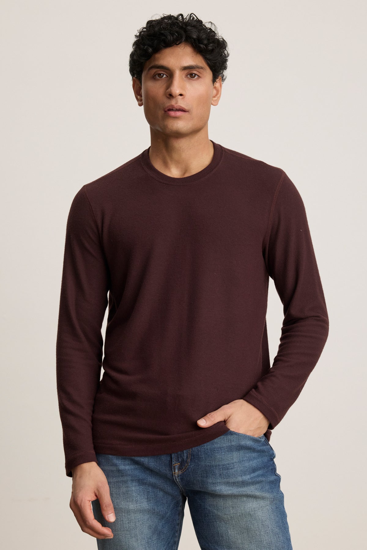 A man with short curly hair wears the Velvet by Graham & Spencer BECKER CREW, a fitted maroon long-sleeve shirt, paired with blue jeans, standing against a plain light background.-39905750745281