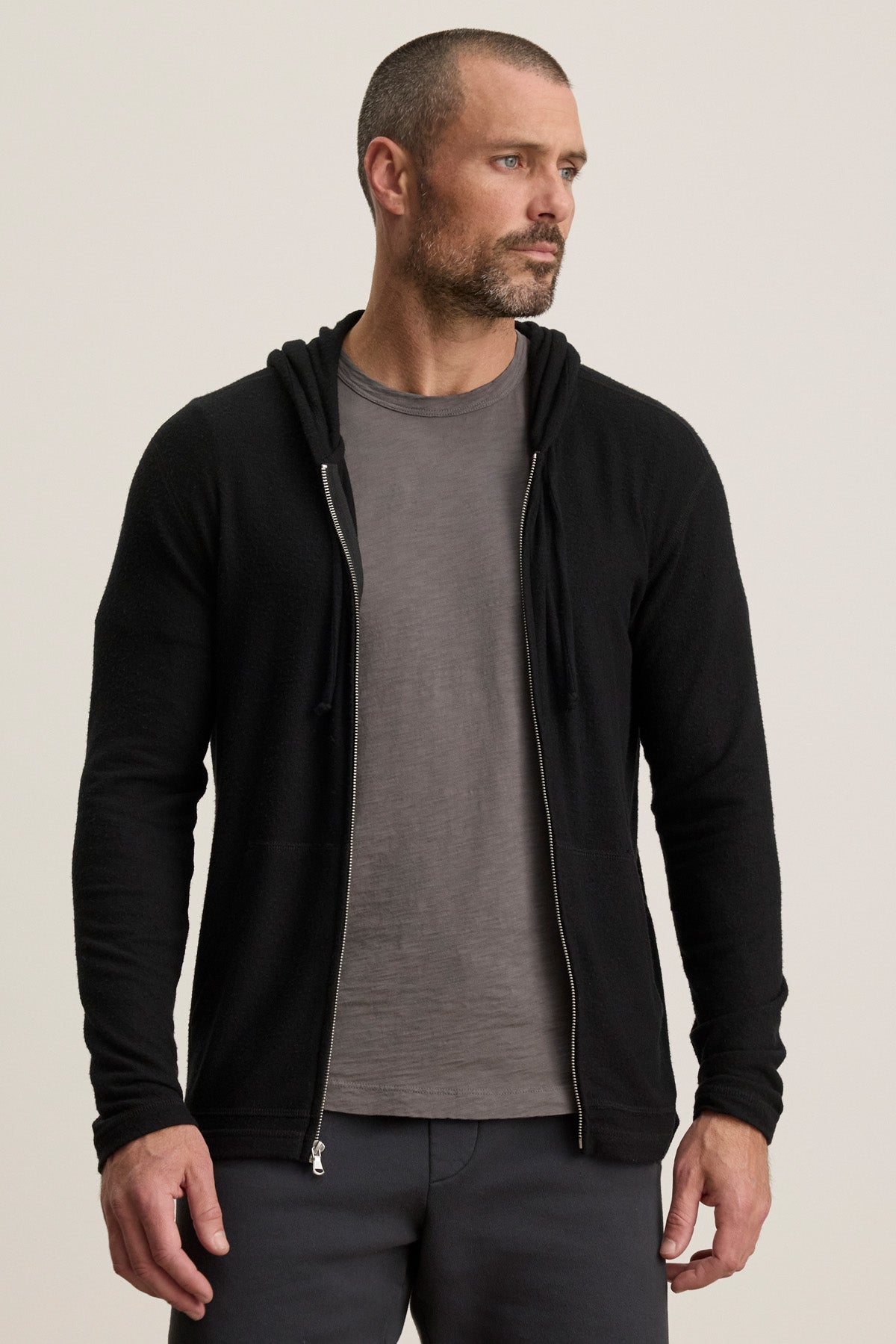 A man with a short beard and cropped hair wears the LOUDON HOODIE by Velvet by Graham & Spencer—a black, relaxed-fit full-zip hoodie—over a gray t-shirt, standing against a plain light background.