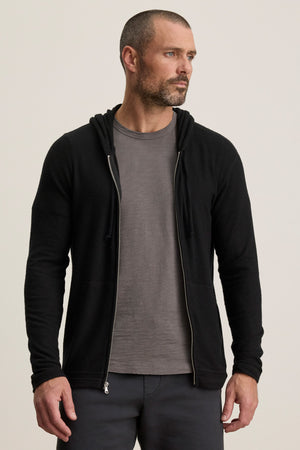 A man with a short beard and cropped hair wears the LOUDON HOODIE by Velvet by Graham & Spencer—a black, relaxed-fit full-zip hoodie—over a gray t-shirt, standing against a plain light background.