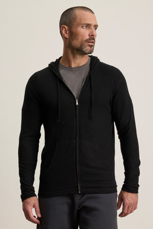 A man wears the LOUDON HOODIE by Velvet by Graham & Spencer in black, layered over a gray t-shirt, standing slightly turned against a plain light background.