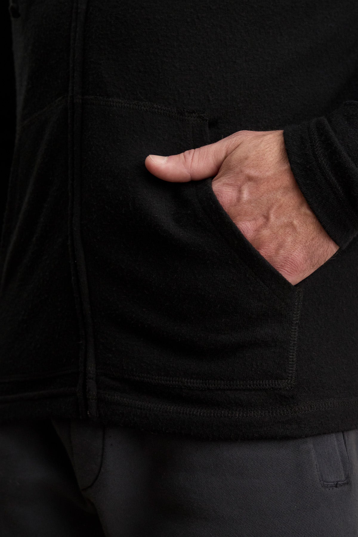 A person’s hand is partially tucked into the front pocket of the LOUDON HOODIE by Velvet by Graham & Spencer—a relaxed fit black fleece jacket—with the arm slightly bent and dark pants visible below.