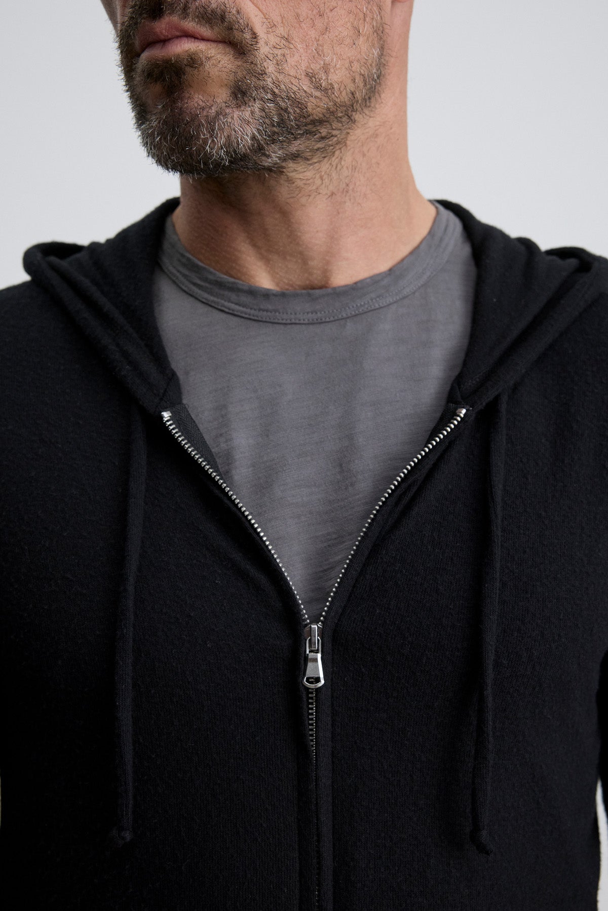 A man with a short beard wears a grey tee and the LOUDON HOODIE by Velvet by Graham & Spencer—a relaxed fit, black full-zip hoodie with drawstrings, partially unzipped. The cozy jersey image is cropped at his nose and chest.