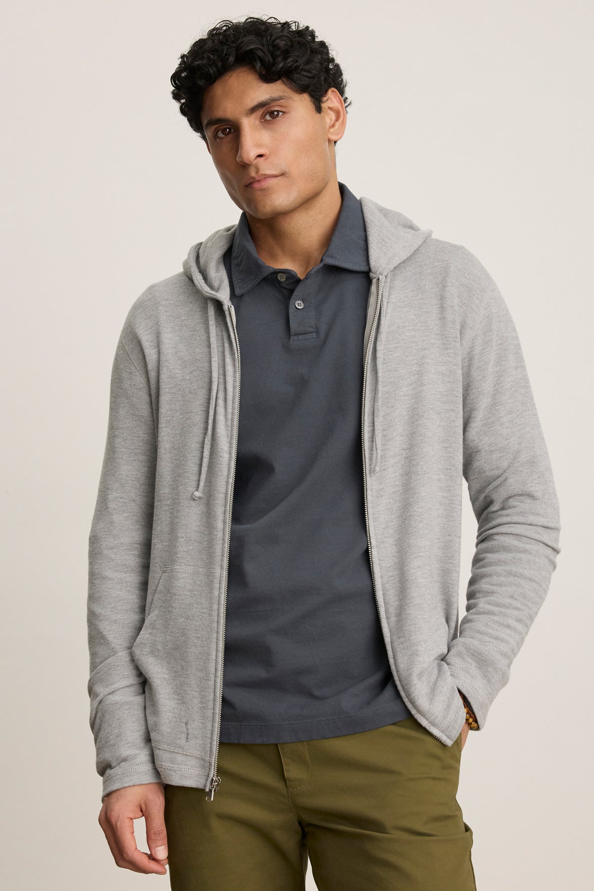 A man stands against a plain background, wearing the LOUDON HOODIE by Velvet by Graham & Spencer—a relaxed fit triblend gray full-zip hoodie—layered over a dark gray polo and olive green pants.-39680474808513
