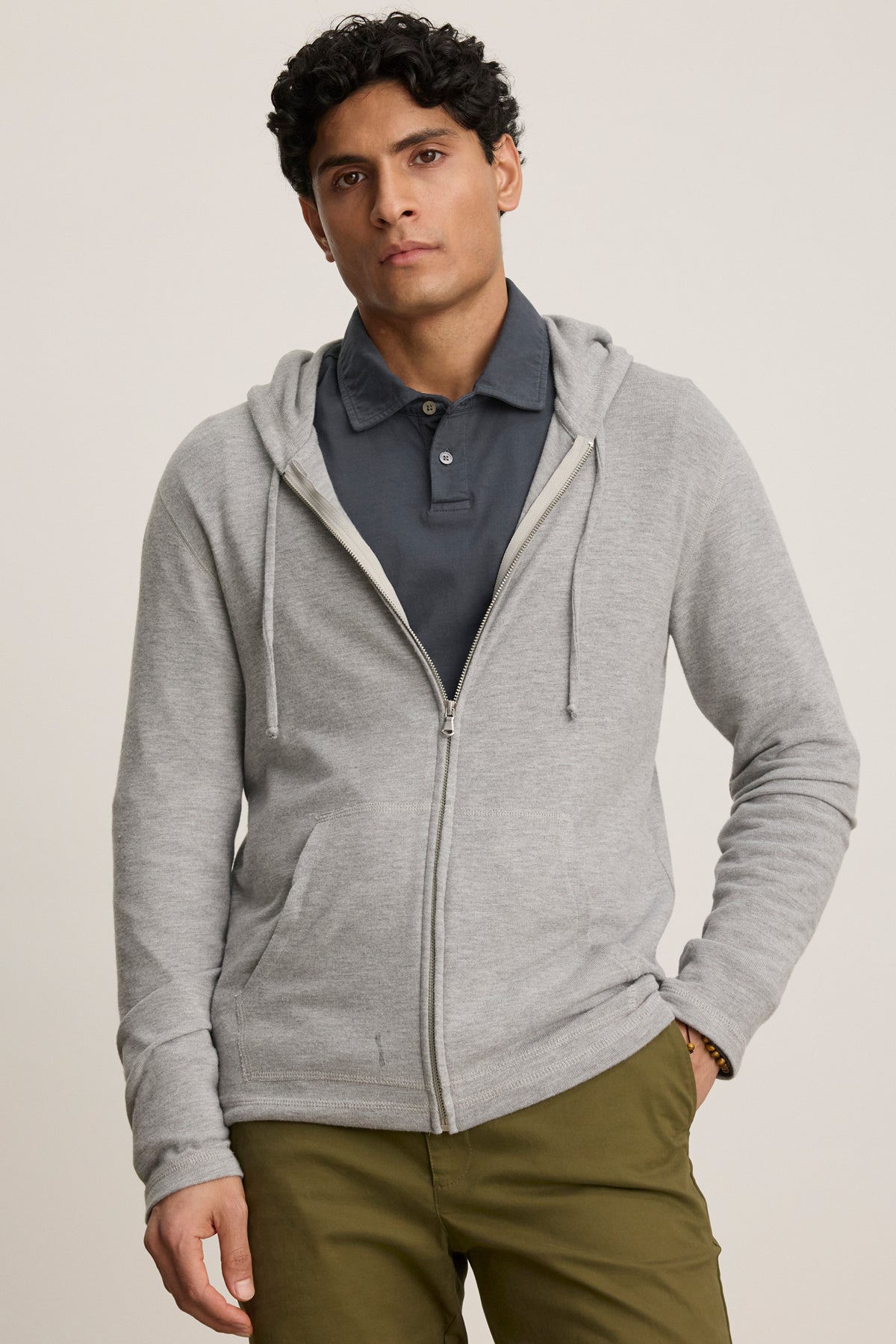   A man wearing the LOUDON HOODIE from Velvet by Graham & Spencer—a relaxed fit, full-zip hoodie in triblend cozy jersey—over a dark grey polo and olive green pants stands against a plain background. 