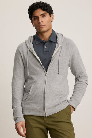 A man wearing the LOUDON HOODIE from Velvet by Graham & Spencer—a relaxed fit, full-zip hoodie in triblend cozy jersey—over a dark grey polo and olive green pants stands against a plain background.