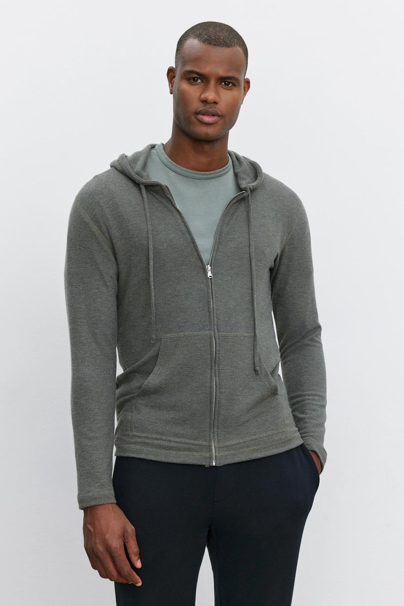 A man dressed in a Velvet by Graham & Spencer LOUDON HOODIE, with a grey shirt and black pants, stands against a plain white background.