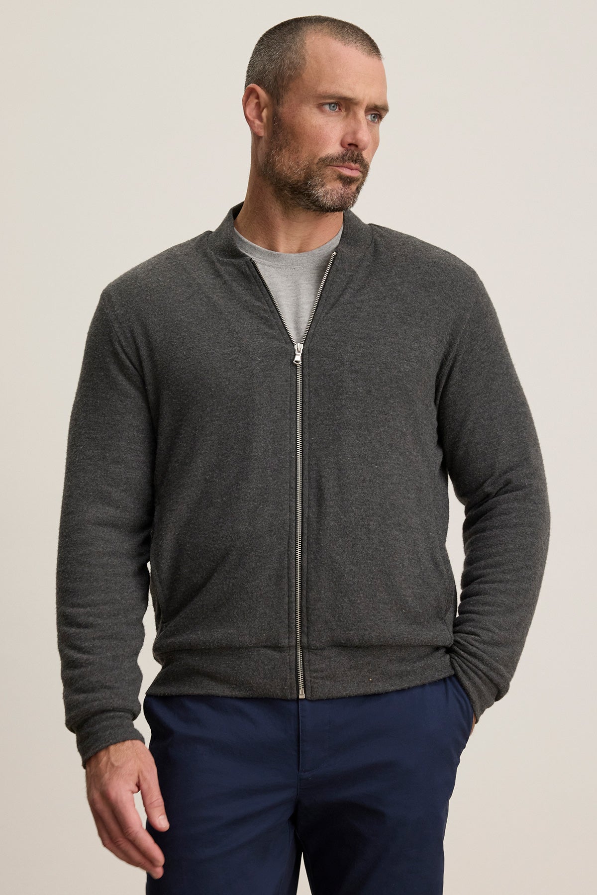 A man wears the MILES JACKET by Velvet by Graham & Spencer over a grey t-shirt and navy blue pants, standing against a plain background.