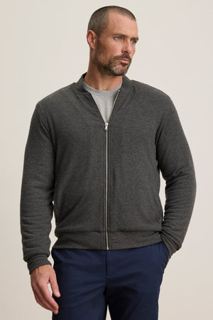 A man wears the MILES JACKET by Velvet by Graham & Spencer over a grey t-shirt and navy blue pants, standing against a plain background.