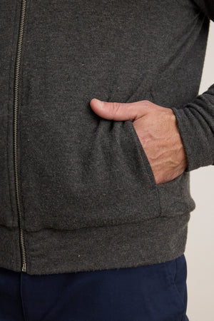 A person wearing the MILES JACKET by Velvet by Graham & Spencer—a dark gray casual layer with a soft knit shell—and navy pants, their right hand tucked into the jacket pocket.