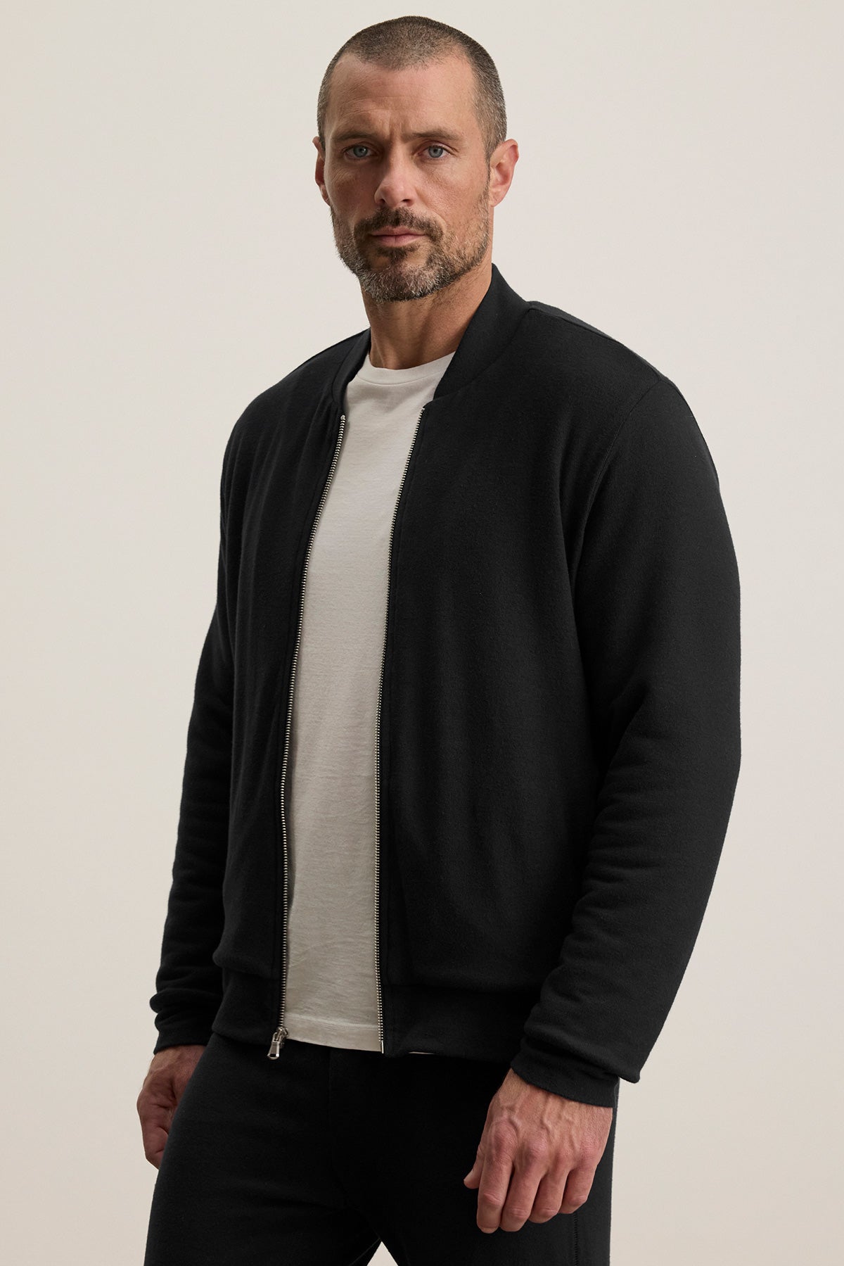 A man with short hair and a beard wears the MILES JACKET by Velvet by Graham & Spencer over a white T-shirt, standing against a plain light background.