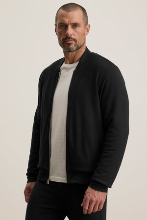 A man with short hair and a beard wears the MILES JACKET by Velvet by Graham & Spencer over a white T-shirt, standing against a plain light background.