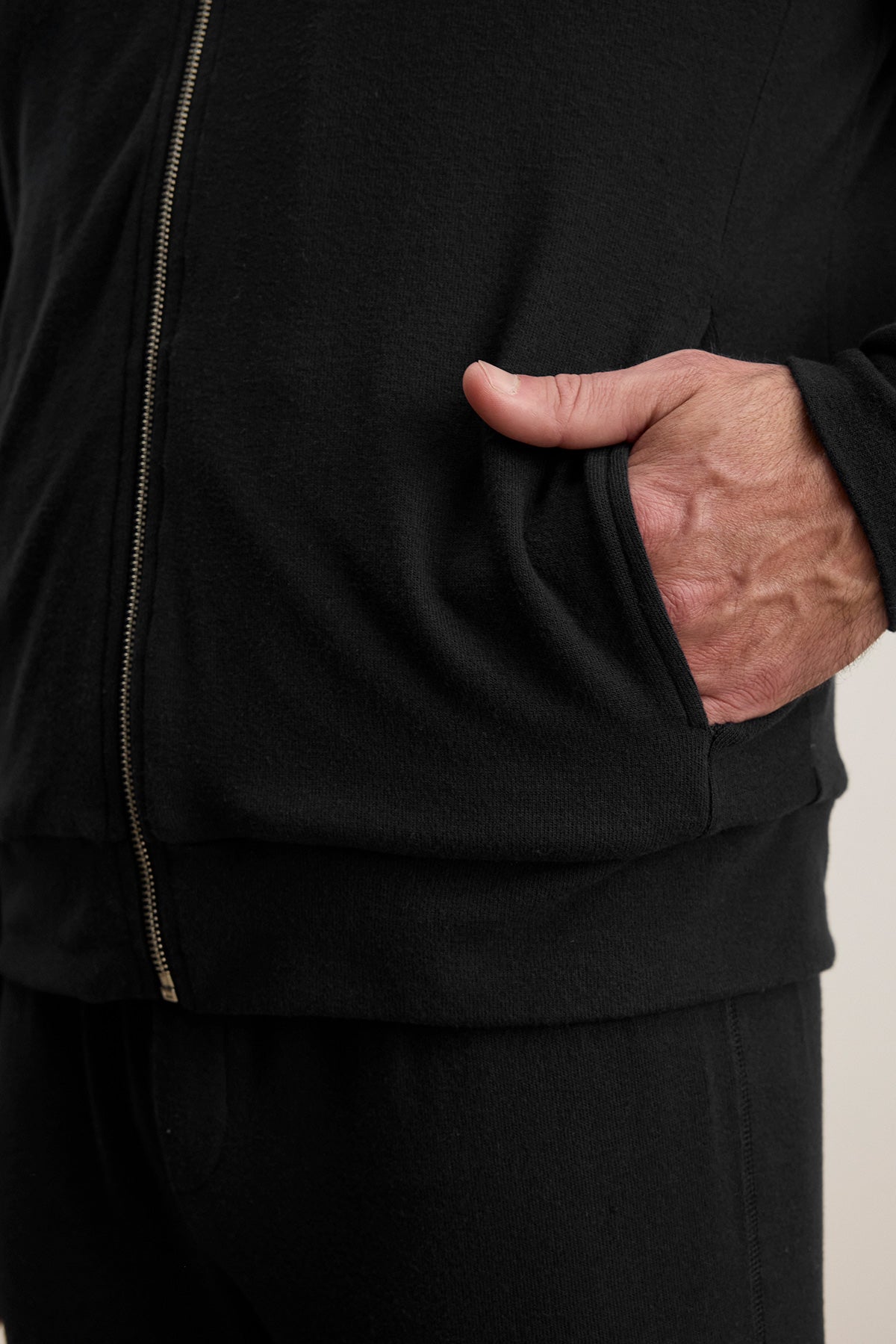 A person wearing the MILES JACKET by Velvet by Graham & Spencer stands confidently with their right hand in the pocket; this black zip-up jacket has a sleek design and pairs well with matching pants.-40307484065985