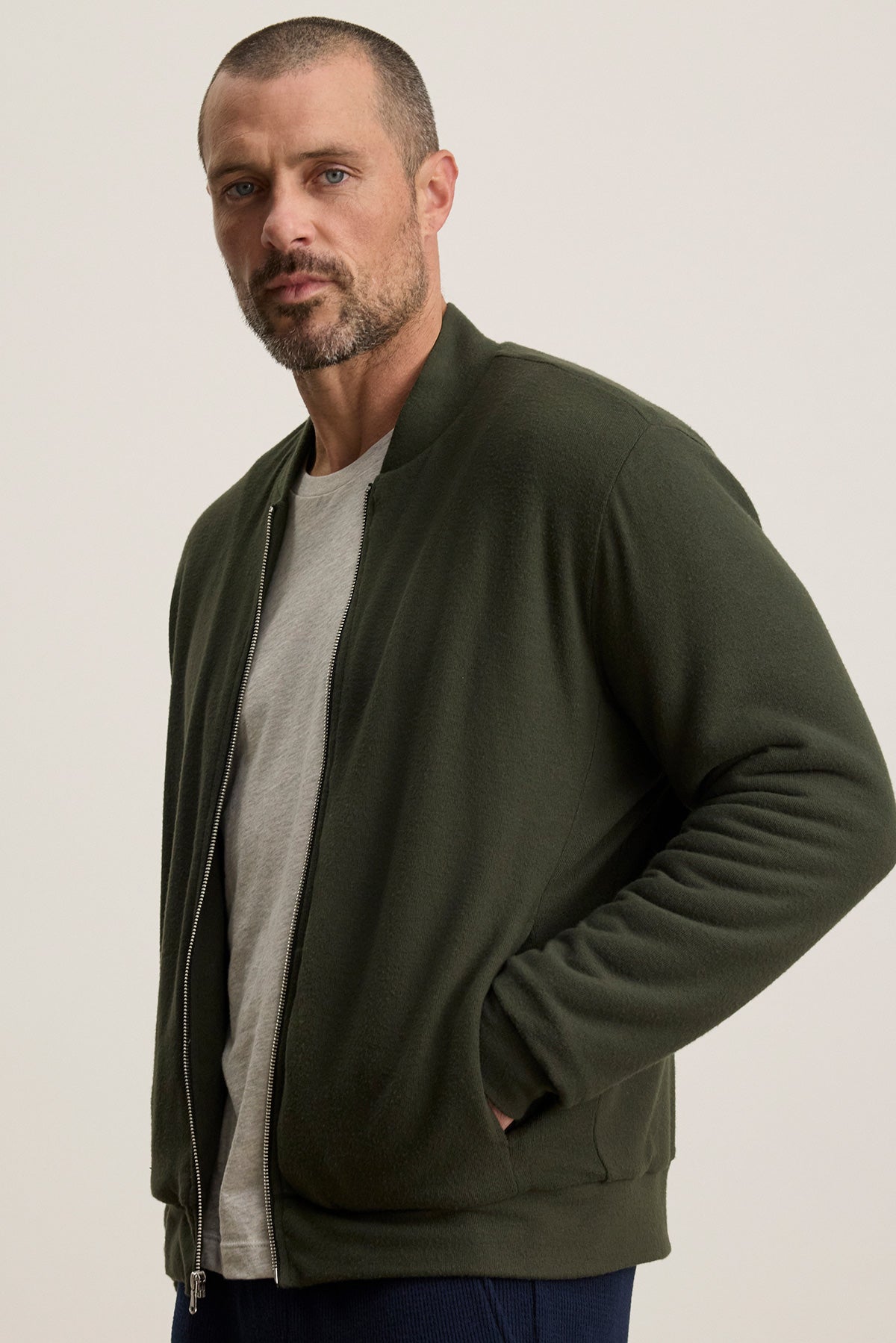 A man with short hair and a beard wears the MILES JACKET by Velvet by Graham & Spencer, a dark green casual jacket layered over a light grey t-shirt, standing with one hand in his pocket against a plain background.-40277366833345