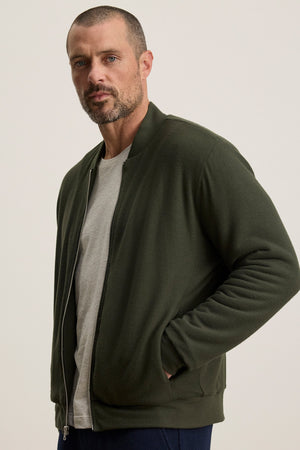 A man with short hair and a beard wears the MILES JACKET by Velvet by Graham & Spencer, a dark green casual jacket layered over a light grey t-shirt, standing with one hand in his pocket against a plain background.