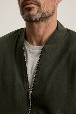 A close-up of a man wearing the MILES JACKET by Velvet by Graham & Spencer in olive green, layered over a light grey t-shirt with the zipper partially open.