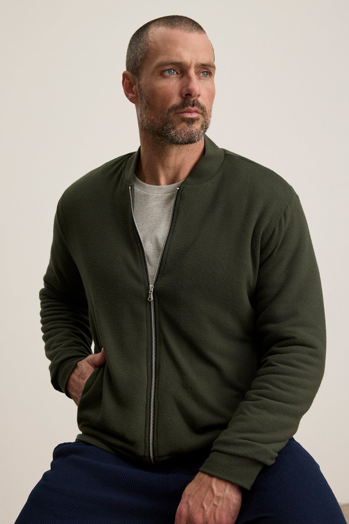 A man with short hair and a beard wears the MILES JACKET by Velvet by Graham & Spencer, an olive green zip-up, over a gray shirt while sitting with one hand on his knee against a plain background.-40277366964417