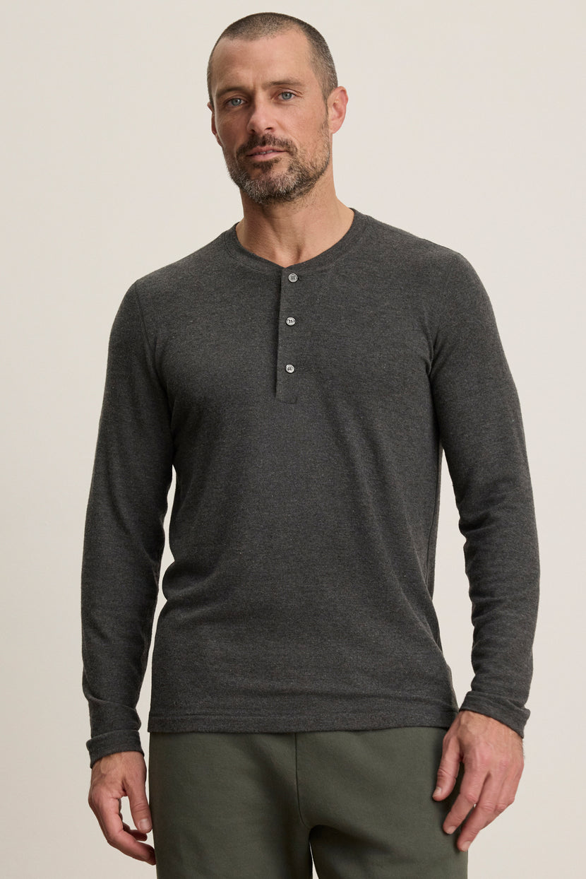 A bearded man wears the RYLAND HENLEY by Velvet by Graham & Spencer in charcoal gray and olive green pants, standing against a plain background—a cozy jersey option for casual occasions.