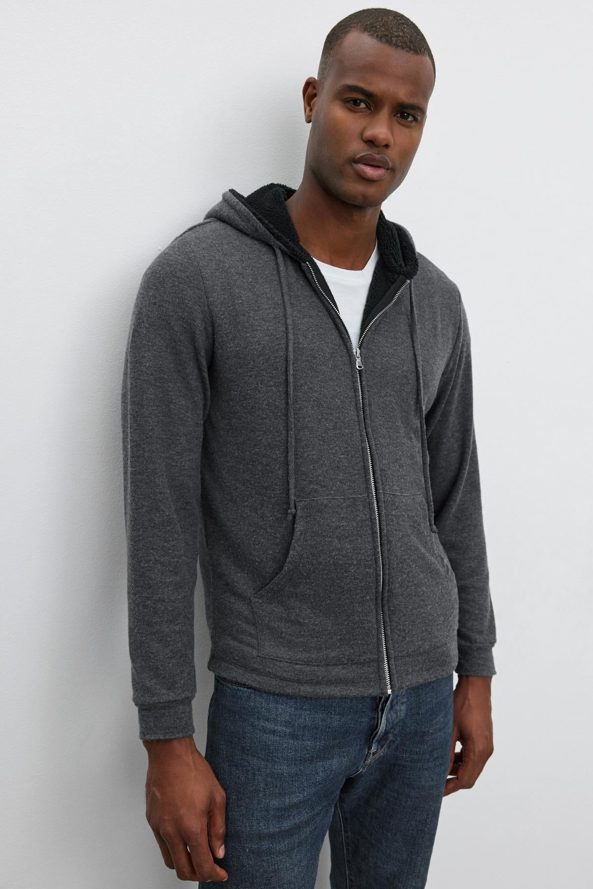   A person stands against a plain background, wearing the Velvet by Graham & Spencer SALVADORE HOODIE in dark gray over a white t-shirt and blue jeans, looking directly at the camera with a neutral expression. The ultra-soft cozy jersey fabric hints at an added layer of comfort. 