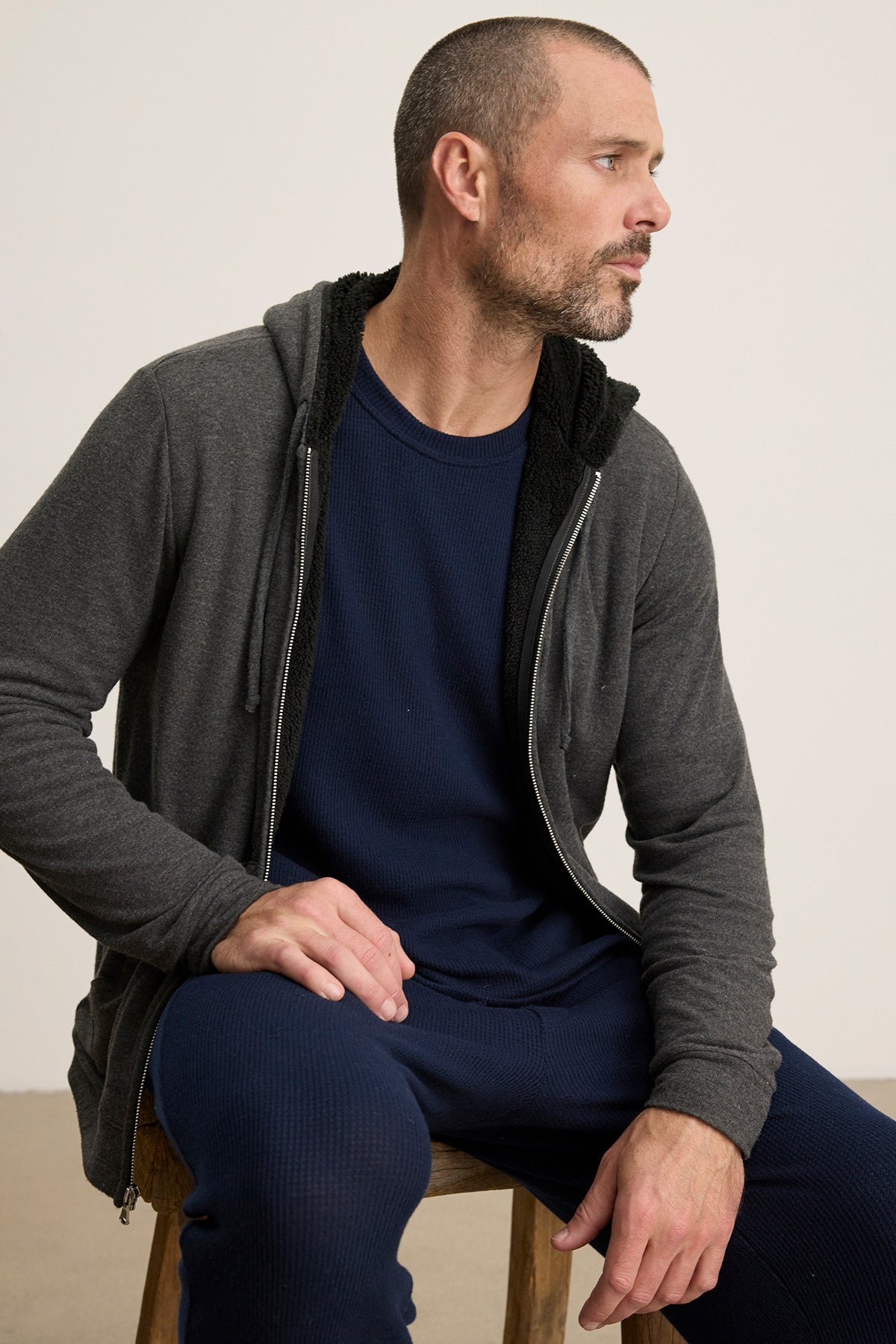 A man with short hair and a beard sits on a wooden stool, wearing the SALVADORE HOODIE by Velvet by Graham & Spencer and navy blue loungewear, looking to the side against a plain background.-40277276655809
