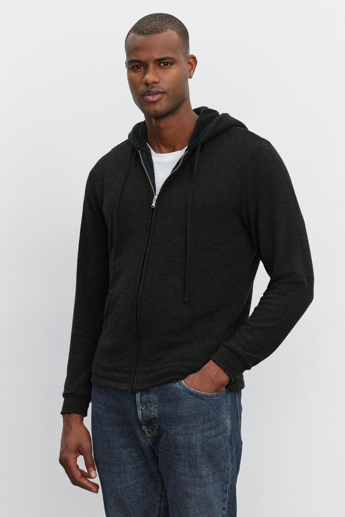   A man stands against a plain background wearing the SALVADORE HOODIE by Velvet by Graham & Spencer over a white t-shirt and blue jeans, with his left hand in his pocket. 