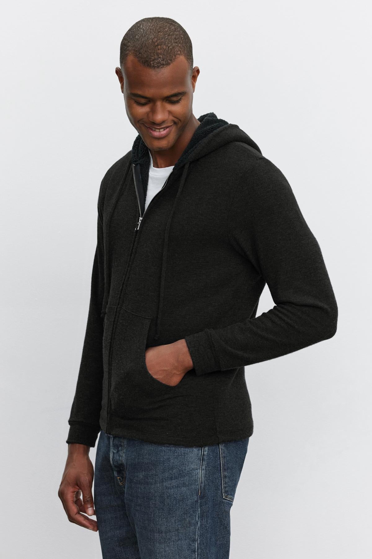   A man wearing a SALVADORE HOODIE by Velvet by Graham & Spencer, complete with faux sherpa lining, stands with one hand in his pocket over a white T-shirt, looking down and smiling. 