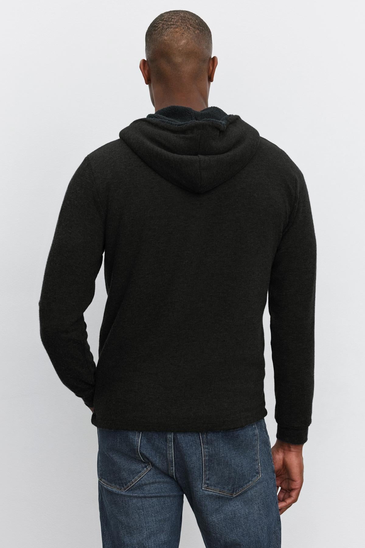   A man is photographed from the back, wearing the SALVADORE HOODIE by Velvet by Graham & Spencer, featuring a black full-zip design with faux sherpa lining, paired with blue jeans. 