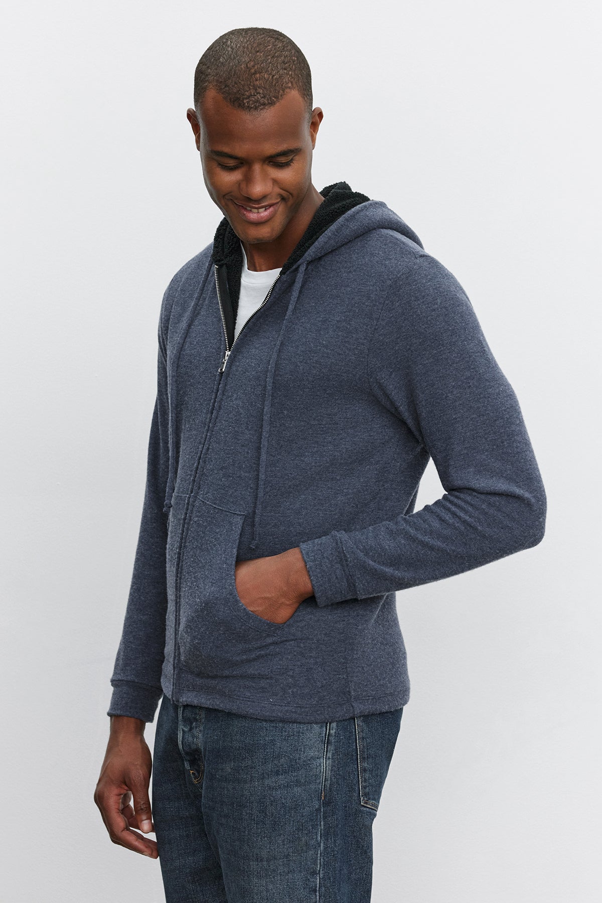 A person wearing the SALVADORE HOODIE by Velvet by Graham & Spencer and blue jeans stands with their hands in their pockets, looking down and smiling.-37893927043265