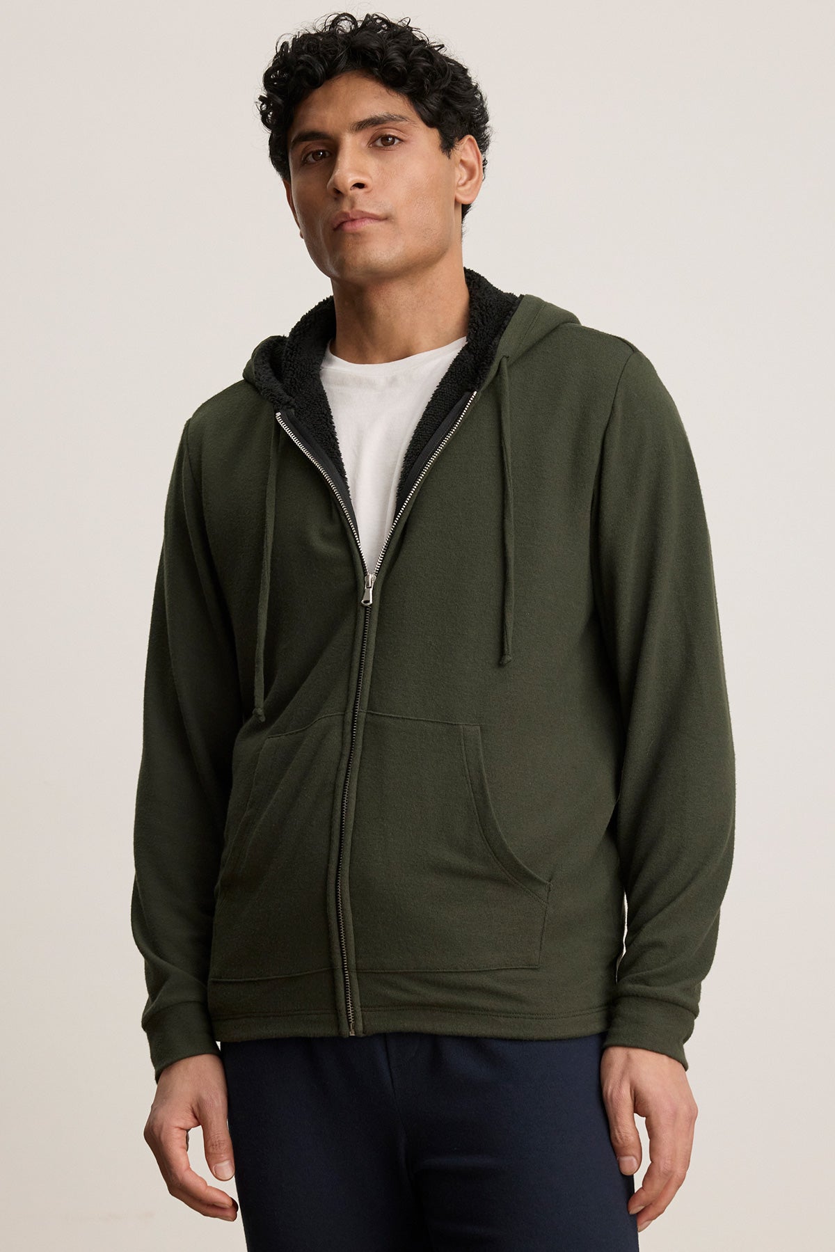 A man wearing the SALVADORE HOODIE by Velvet by Graham & Spencer, a dark green zip-up hoodie with an adjustable drawstring hood, over a white T-shirt and dark pants stands against a plain light background.-40277251424449