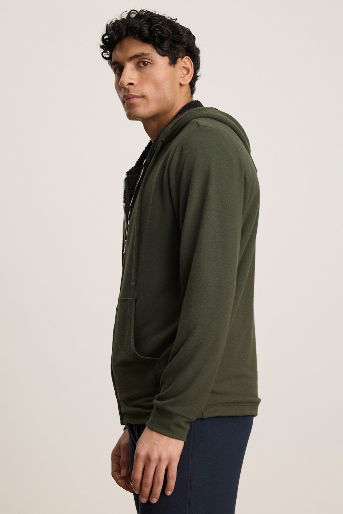 A man stands in profile wearing the Velvet by Graham & Spencer SALVADORE HOODIE, a green zip-up with adjustable drawstring hood, paired with dark pants against a plain light background.-40277251457217
