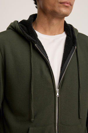 A person wearing the Velvet by Graham & Spencer SALVADORE HOODIE, a dark green zip-up with black fleece lining and a white t-shirt underneath; only their lower face and torso are shown.