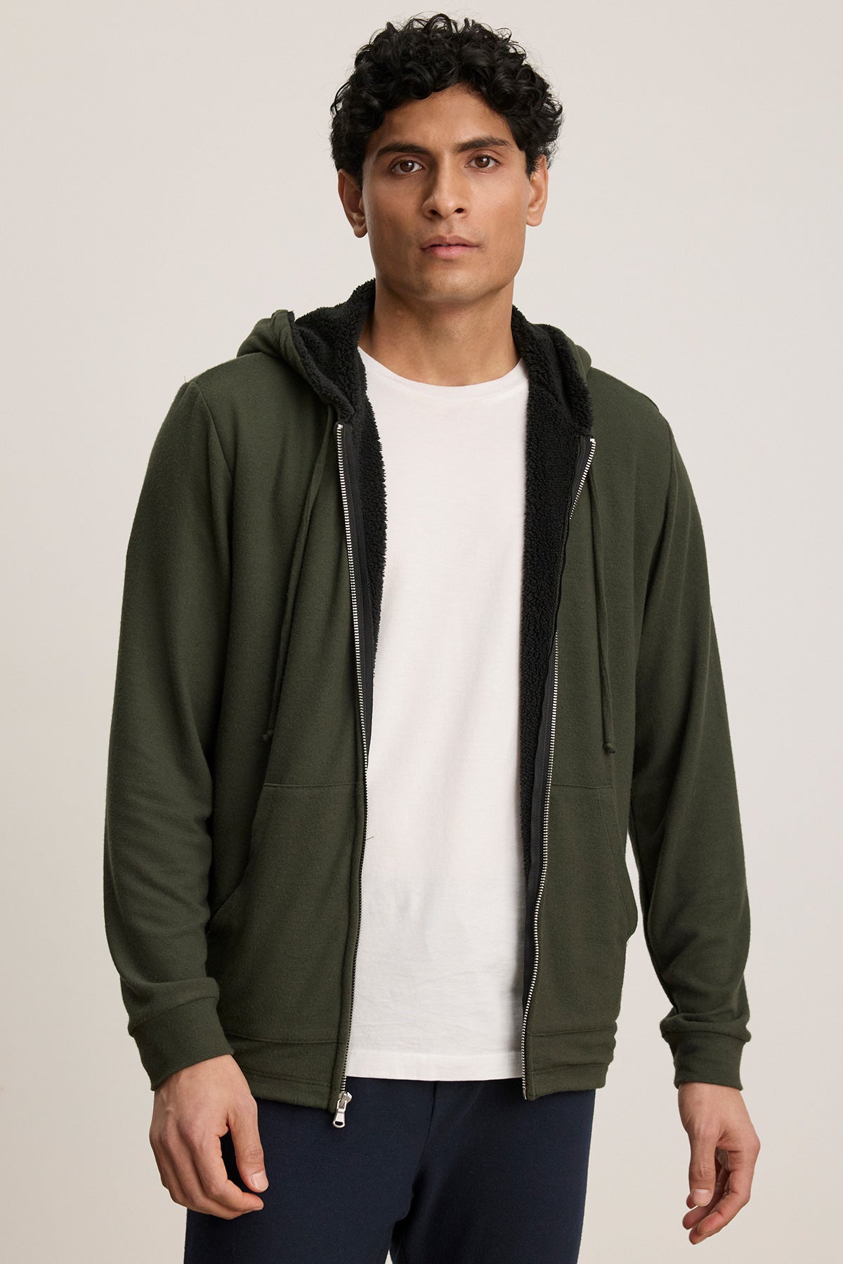 A person wears the SALVADORE HOODIE by Velvet by Graham & Spencer—a dark green zip-up with faux shearling lining and an adjustable drawstring hood—over a white t-shirt, standing against a plain light background.