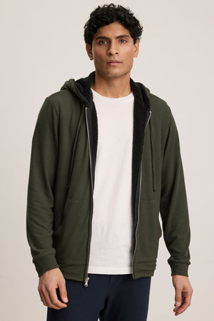 A person wears the SALVADORE HOODIE by Velvet by Graham & Spencer—a dark green zip-up with faux shearling lining and an adjustable drawstring hood—over a white t-shirt, standing against a plain light background.