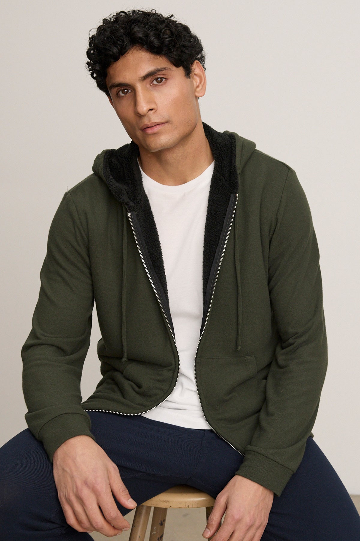 A person with short curly hair wears the SALVADORE HOODIE by Velvet by Graham & Spencer, a dark green zip-up with adjustable drawstring hood, over a white tee and dark pants while sitting on a wooden stool against a plain background.